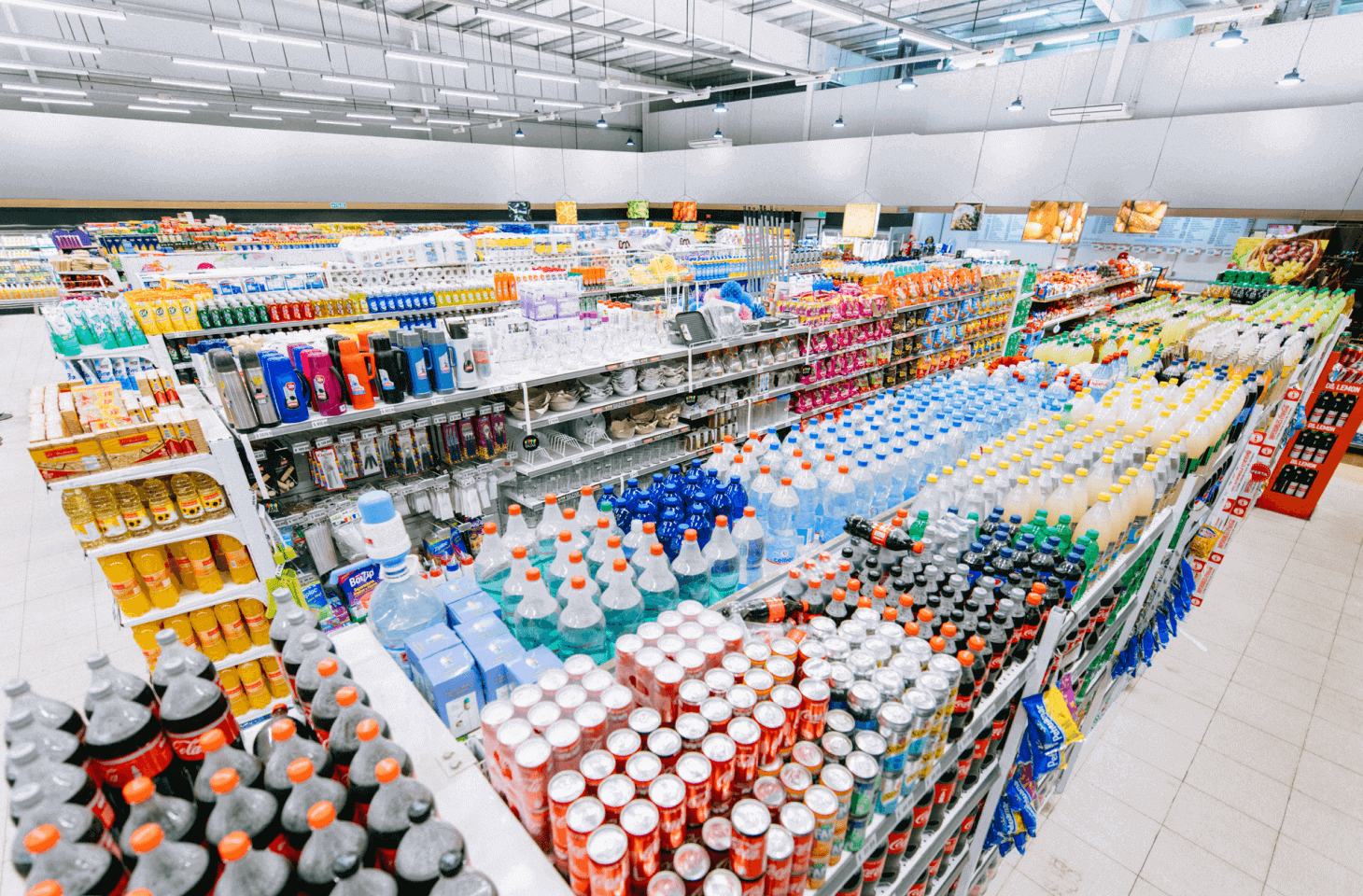 Wide view of a grocery store aisle with shelves stocked with various beverages, cleaning supplies, and household items under bright lighting.